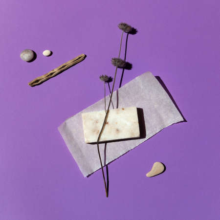 A bar of natural organic handmade cream-colored soap on a beautiful violet background surrounded by sea stones and dry herbs, close-up, top view. Abstraction, minimalism.の写真素材