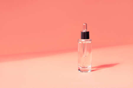 A beautiful transparent bottle with a cosmetic moisturizing, anti-aging serum made from natural ingredients on a coral paper background in the sun with a beautiful shadow. Front view, place for text.の写真素材