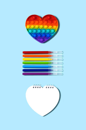 Back to school concept. Popular silicone anti-stress push pop fidget toy pop it rainbow colors, felt-tip pens and notebook in the form heart close-up, on a blue background vertical, top view.の写真素材