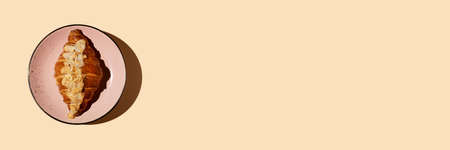 Banner freshly baked buttery croissant with almond frangipan sprinkled with nut petals on a pink plate on a beige background with hard shadow, copy space. Coffee, pastry shop breakfast concept.の写真素材