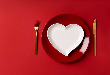 Festive table setting for valentine's day or wedding anniversary red plate and white heart shaped plate with golden cutlery on red background. Place for an inscription as a invitation card or menu.の写真素材