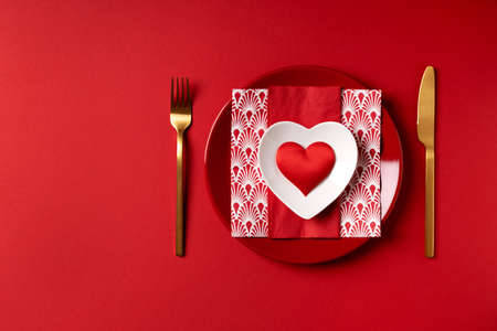 Festive table setting for valentine's day or wedding anniversary red plate with a heart on a napkin and golden cutlery on a red background. Place for an inscription as a layout for an invitation card.の写真素材