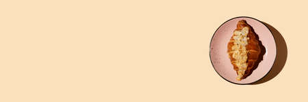 Banner freshly baked buttery croissant with frangipan almond sprinkled with nut petals on a pink plate on a beige background with hard shadow, copy space. Coffee, pastry shop breakfast concept.の写真素材