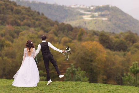 Back view of a young amazing groom and bride holding hands and looking at the mountains.の写真素材