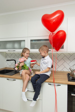 A boy and a girl are sitting on a kitchen with a bouquet of pink tulips and red gift box. Love, friendship and funの写真素材