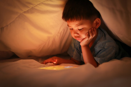 Little asian boy attention on online device while lying under white blanket in the bedroom at night, bright screen light reflex on kid face. Learning concepts of internet and technology for children.の写真素材