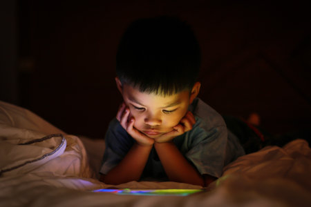 Little asian kid alone watching tablet device, lying on white duvet bed with chin on hands, in background darkness bedroom night time. Colourful bright light from screen reflex on the boy face.の写真素材