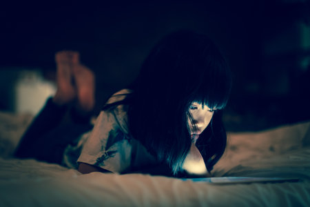 Little child using tablet in darkness bedroom while lying on the bed, bright light screen reflex on her face.  Concepts of social network online issue and kids permission to access internet.の写真素材