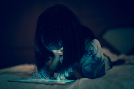 Asian child face down looking at tablet screen while lying on bed, in darknss bedroom background, concepts of cyberbullying and social media safety permisstion for kids and teenagers.の写真素材