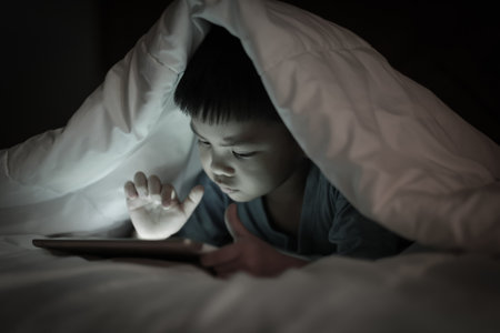 Asian boy using tablet screen while lying on bed under white blanket, in the bedroom at night,...