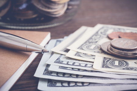 Money banknote US dollar and coins put on wooden table with silver pen, notebook and saving jar on background, vintage style. Currency, financial economy, earning income, budget management conceptsの写真素材