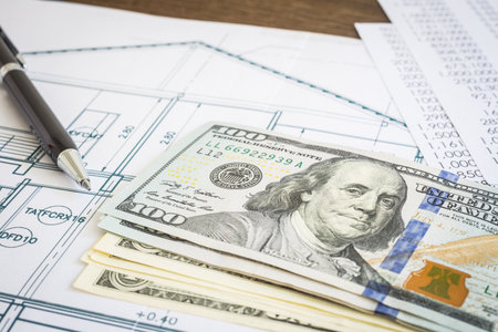 New dollar banknotes or money, house blueprint floor plan, paperwork and black pen put on wooden table. Concepts of construction budget, mortgage, home loan and refinance.の写真素材
