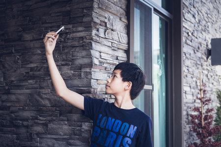 Asian boy teenager in dark blue t-shirt with alphabet word pattern, raises arm up and using mobile phone to take photo selfie his picture. Daylight outdoor background with stone wall, window, house.の写真素材