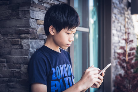 Asian boy teenager in dark blue t-shirt using wireless smartphone for reading informantion and send message outside the home. Daylight outdoor background with stone wall, window, house.の写真素材