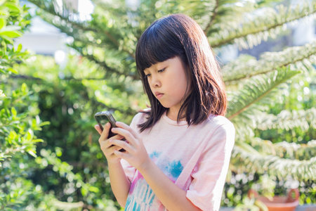 Little cute asian kid girl in pink t-shirt looking at device and using wireless smartphone send message,  play game and internect. Daylight outdoor  garden trees, blur in background.の写真素材