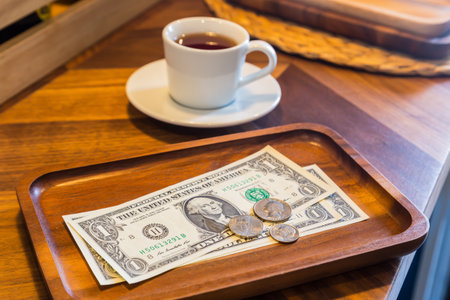 Dollar banknotes and US coins on wooden tray, on coffee shop table with a white cup of tea on background. Dim light room, modern and minimal style. Concepts for customer service, checking, money tips.の写真素材