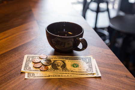 Dollar banknotes and US coins on wooden cafe's table and a black cup of tea or coffee. Dim light room, modern and minimal style. Concepts for fee charge, customer service, bill checking, money tips.の写真素材