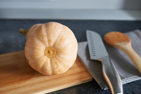 Pumpkin on a cutting board with a knife and spoon on a kitchen countertop.の写真素材