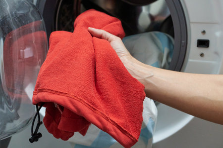 A woman's hand puts clothes into a washing machine. A red shirt in a woman's hand against the background of a washing machine.の写真素材
