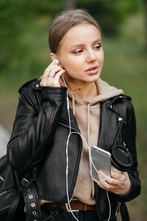 A beautiful woman in a leather jacket walks through the park, inserts headphones into her ears to listen to musicの写真素材