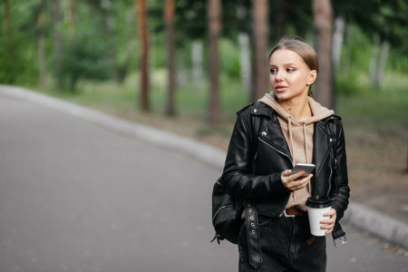A beautiful woman in a leather jacket walks in the park and smiles pretty while looking to the side with a phoneの写真素材