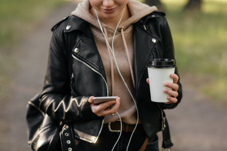A woman with headphones in her ears holds the phone in her hand and looks into it. Walking in the park.の写真素材