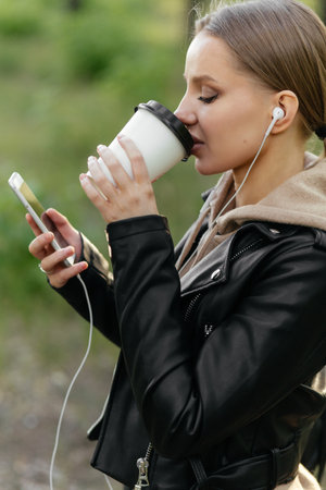 A beautiful woman in headphones walks through the park, talks on a video call from a phone and drinks coffee.の写真素材