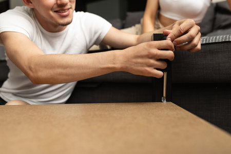 man is screwing a screw into the table with a screwdriver. a beautiful woman sits on the sofa beside him and watchesの写真素材