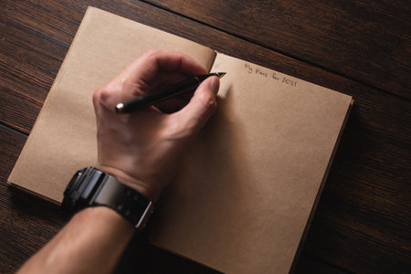 A man writes in a notebook My plans for 2021 with a pen on a dark wooden table.