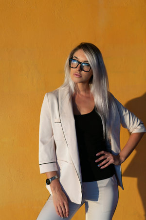 Beautiful blonde in a beige trouser suit and glasses against the background of an orange wallの写真素材