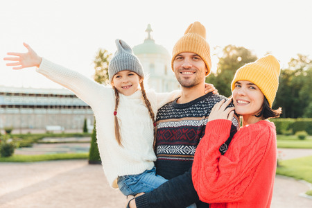 Cute lovely girl being on father`s hands, wave her hand as sees something wonderful into distance, spends her free time with father and mother, pose outdoors. Family embrace each other with great loveの写真素材