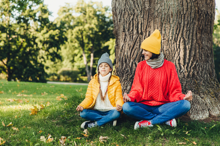 Body language concept. Carefree little kid sits near her beautiful mother, keep hands in mudra sign, keep eyes closed, try to calm down after hard work at home, admire fresh air in beautiful parkの写真素材