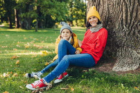 Outdoor portrait of beautiful female dressed in warm cap and warm loose sweater spend weekends together with her little daughter, sit on green grass near tree, enjoy fresh air and wonderful natureの写真素材