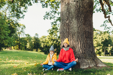 Yoga and meditation concept. Beautiful young woman in knitted clothes and small child keep eyes closed, meditate outdoors in green park, feel relaxed, calm and peaceful, keep hands in mudra signの写真素材