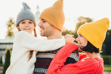 People and relationship concept. Family have unforgettable time together, embrae each other, wear trendy knitted hats. Adorable small girl with pigtails embrace her father, looks happily into cameraの写真素材