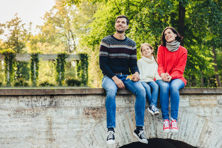 Horizontal portrait of adorable child sits between her mother and father, enjoy beaitiful landscapes from top of ancient bridge, breath fresh air, look into distance. Family and relationship conceptの写真素材
