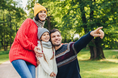 Three family members spend time together, look at beautiful lake in park, indicate with fingers, being in good mood, smile pleasantly. Father, mother and daughter enjoy togetherness, calm atmosphereの写真素材