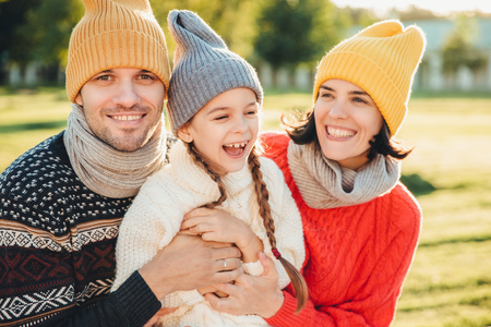 Playful little child with pigtails wears warm clothes, spends free time with lovely affectionate parents, have happy expression, feel relaxed. Three young family members enjoy pleasant momentsの写真素材