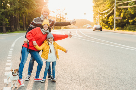 Portrait of family: mother, father and little daughter walk with pet, stand on road, raise thumbs as show ok sign,express approval or agreement, have good mood after spending time on fresh airの写真素材