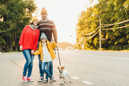 Happy family wear warm clothes walk with dog on road, stand close to each other as pose in camera. Little girl shows ok sign as being glad to spend free time with her parents and favourite petの写真素材