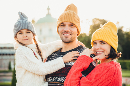 Outdoor portrait of friendly family stand close to each other, have broad smiles. Pretty woman in yellow trendy hat and red sweater, handsome young man holds little adorable daughter on handsの写真素材