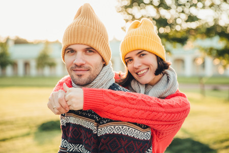 Smiling glad young female in hat and warm cotton sweater embraces her husband who stands back, enjoy spending weekends together, have walk in garden or park, admire splendid sunny autumn weatherの写真素材