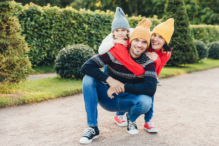 Beautiful female, little girl embrace their handsome father and husband, have good relationship, have active lifestyle, pose against green trees. Family have walk together, embrace each otherの写真素材