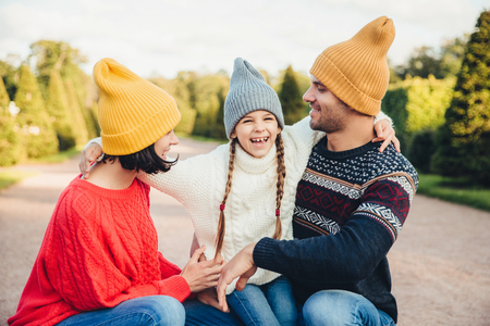 Happy small girl embrace her mother and father, have fun together, walk in beautiful park or on road. Parents look at her lovely daughter, try to do best for her. Happy family have good rest outdoorsの写真素材