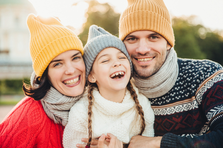 Horizontal portrait of smiling happy woman and man have good time with their daughter, wear knitted hats, warm sweaters and scarfs, going in park to have walk together. Spending time in family circleの写真素材