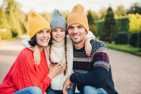 Outdoor portait of beautiful brunette female, handsome man, pretty small girl embrace together, have pleasant mood, walk in park, enjoy calm atmosphere and splendid sunny weather. Friendly familyの写真素材
