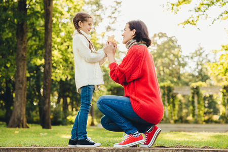 Young mother in warm knitted red sweater plays with her small daughter in park, gives her leaf, enjoy sunny autumn weather.の写真素材