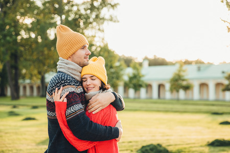 People and closeness concept. Young couple in love have date, embrace each other, feel support, being alone in park, have perfect relationships. Handsome man in warm sweater and hat hugs girlfriendの写真素材