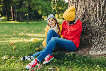 Funny little child and her mother sit together near tree, have walk in autumn park, enjoy wonderful unforgettable time together. Friendly mother and small girl, relax outdoor. Relaxation conceptの写真素材