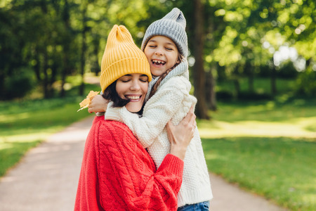 Parenthood, happiness and lifestyle concept. Beautiful female and her daughter have warm embrace, dressed in knitted clothes, have walk during autumn day, enjoy sunny weather, love each otherの写真素材